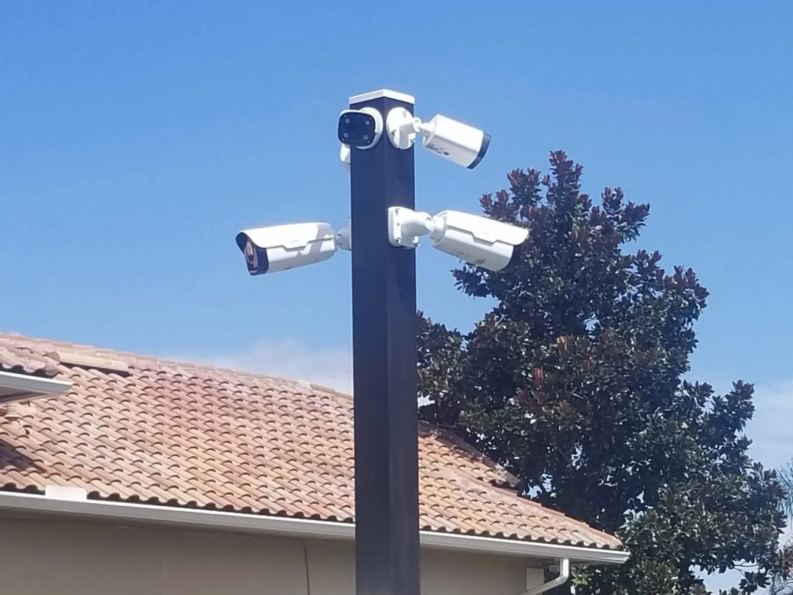 Four outdoor bullet cameras on a pole at a gated property entrance, aimed to cover every approach, with a tile-roofed house behind